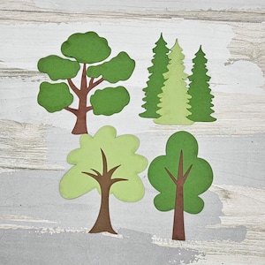 May include: Four paper cut-out trees in varying shades of green and brown. The trees include a large oak, three evergreen trees, and two other deciduous trees. The trees are arranged on a light gray and white wooden background.