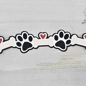 May include: A decorative dog-themed design featuring two black paw prints and two bone shapes, each with a red heart. The design has a black outline and is set against a light gray background.
