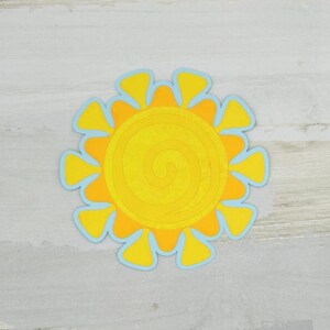 May include: A bright yellow sun-shaped decoration with a spiral center and yellow rays outlined in light blue. The sun is centered on a light gray wooden surface.