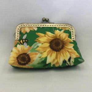 May include: A green fabric coin purse with a gold metal frame. The purse is decorated with a sunflower and bee pattern.