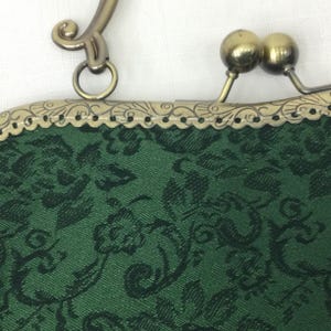May include: Close-up of a green fabric purse with a black floral pattern. The purse has a decorative metal frame with a clasp and handle. The metal has a gold finish.