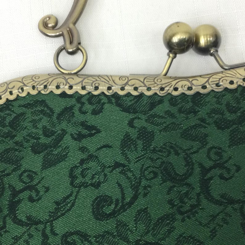 May include: Close-up of a green fabric purse with a black floral pattern. The purse has a decorative metal frame with a clasp and handle. The metal has a gold finish.