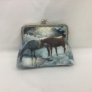 May include: A silver-framed coin purse featuring a serene winter scene with two horses. One horse drinks from a stream, while the other stands nearby. The purse has a clasp closure and a fabric exterior.