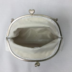 Puede incluir: Clutch de sat&eacute;n blanco con marco plateado y cierres en forma de coraz&oacute;n. La cartera est&aacute; abierta y el interior est&aacute; forrado con sat&eacute;n blanco.