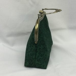 May include: A dark green clutch with a floral pattern. The bag has a metal clasp and a handle. The clasp and handle are a brass color. The bag is made of a textured fabric.