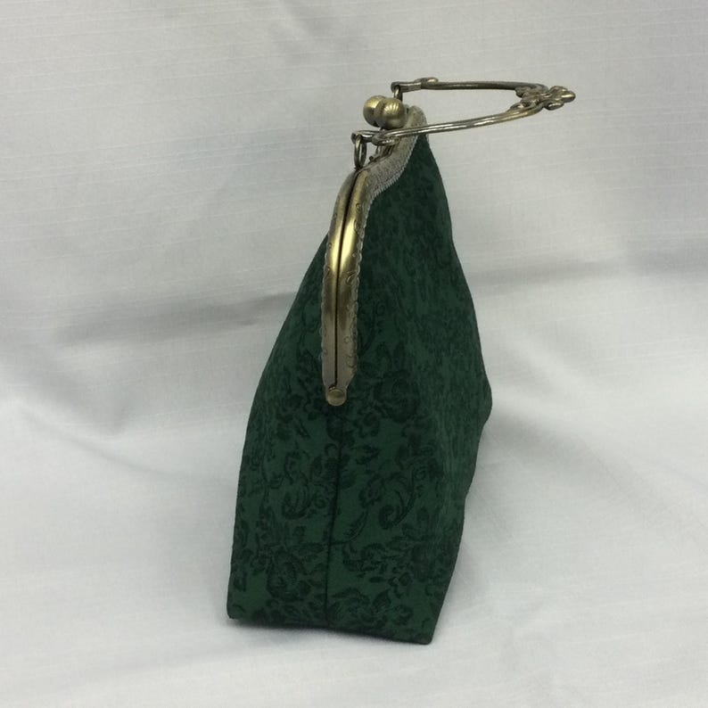 May include: A dark green clutch with a floral pattern. The bag has a metal clasp and a handle. The clasp and handle are a brass color. The bag is made of a textured fabric.