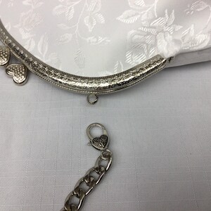 Puede incluir: Una cartera de monedas de sat&eacute;n blanco con un marco de metal plateado y una cadena con un cierre en forma de coraz&oacute;n. La cartera tiene un patr&oacute;n floral y est&aacute; decorada con corazones plateados.