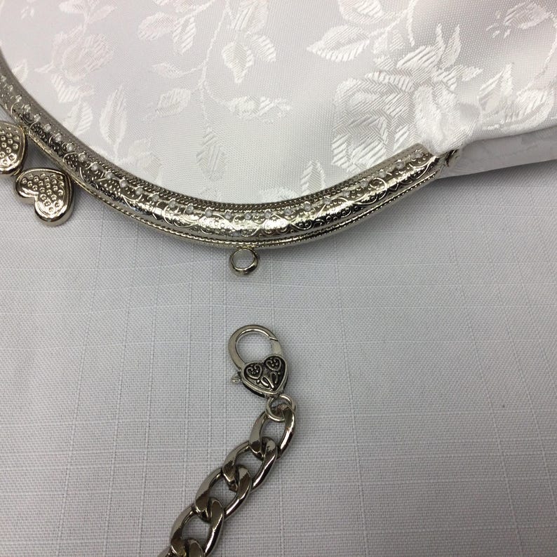 Puede incluir: Una cartera de monedas de sat&eacute;n blanco con un marco de metal plateado y una cadena con un cierre en forma de coraz&oacute;n. La cartera tiene un patr&oacute;n floral y est&aacute; decorada con corazones plateados.