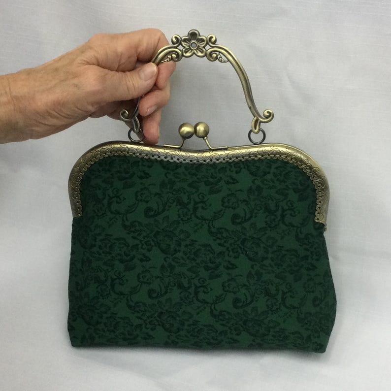 May include: A dark green handbag with a floral pattern. The bag has a decorative bronze-colored frame and handle. The handle features a floral design. The bag is held by a hand.