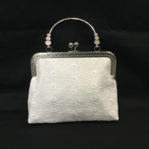 May include: A white handbag featuring an intricate floral eyelet embroidery pattern. The bag has a silver-toned metal frame with a kiss-lock closure and a semi-circular handle adorned with small, clear, sparkling crystals on each side.