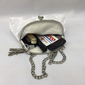 Puede incluir: Un bolso de mano blanco de sat&eacute;n con un estampado floral y una correa de cadena plateada. El bolso est&aacute; abierto y contiene llaves, un l&aacute;piz labial, un tel&eacute;fono y una tarjeta de cr&eacute;dito.
