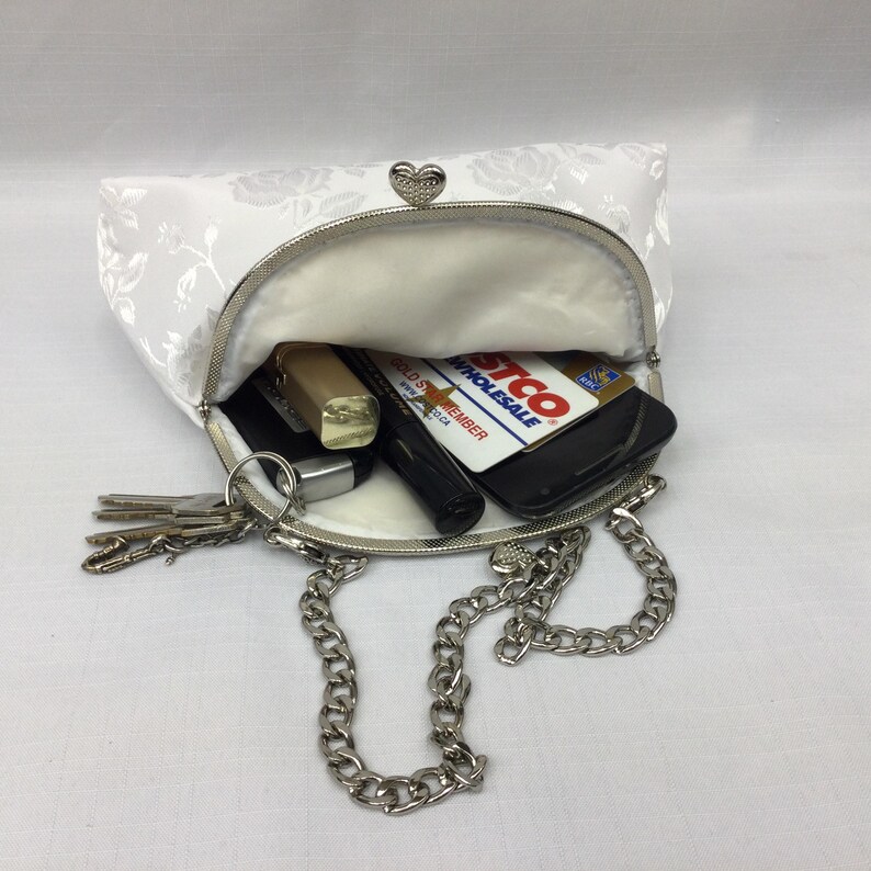 Puede incluir: Un bolso de mano blanco de sat&eacute;n con un estampado floral y una correa de cadena plateada. El bolso est&aacute; abierto y contiene llaves, un l&aacute;piz labial, un tel&eacute;fono y una tarjeta de cr&eacute;dito.