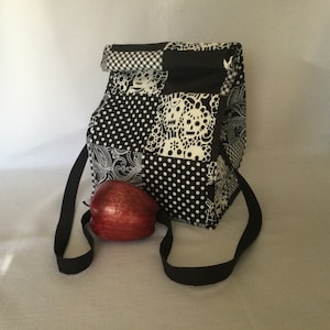 May include: Black and white patterned lunch bag with a black strap. The bag has a patchwork design with skulls, polka dots, and paisley patterns.