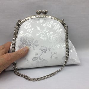 Puede incluir: Un bolso de mano de sat&eacute;n blanco con una correa de cadena plateada y un cierre de coraz&oacute;n plateado. El bolso tiene un patr&oacute;n floral.