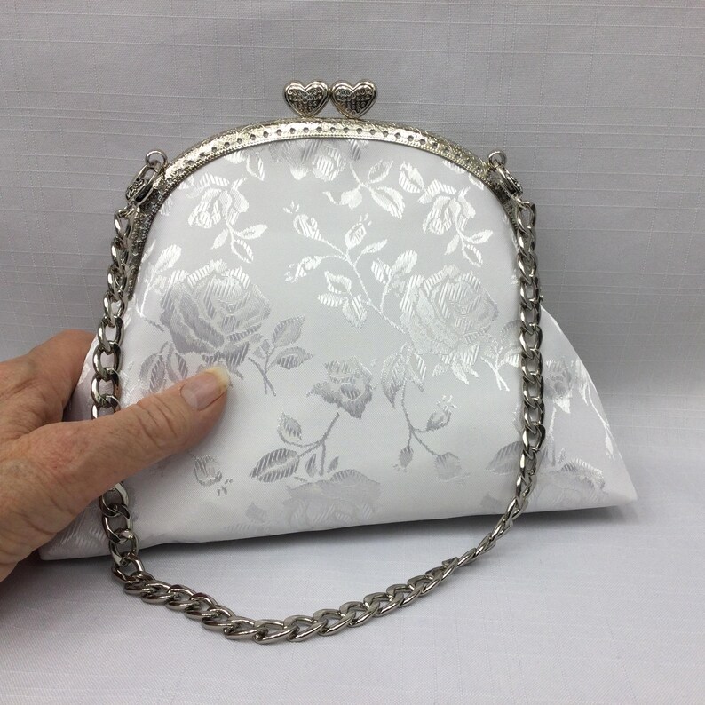 Puede incluir: Un bolso de mano de sat&eacute;n blanco con una correa de cadena plateada y un cierre de coraz&oacute;n plateado. El bolso tiene un patr&oacute;n floral.