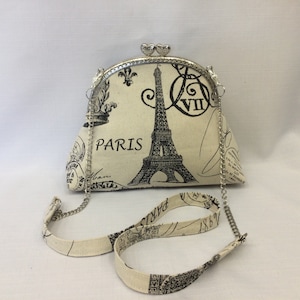 May include: Cream-coloured handbag with a silver clasp and chain strap. The bag features a black Eiffel Tower print with the word "PARIS" and other Parisian-themed graphics. The clasp is adorned with two heart-shaped details.