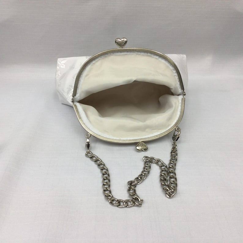 Puede incluir: Una cartera de monedas de sat&eacute;n blanco con una correa de cadena plateada y cierres en forma de coraz&oacute;n.