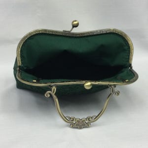 May include: Open, dark green clutch purse with a brass-colored clasp and handle. The interior is a matching dark green fabric. The clasp is a ball shape, and the handle has a floral design. The purse is open, showing the interior.