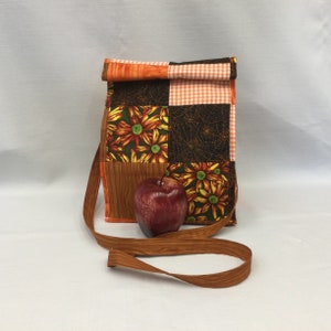 May include: A brown and orange patchwork lunch bag with a long brown strap. The bag has a checkered pattern, a floral pattern, and a spiderweb pattern. A red apple is in front of the bag.