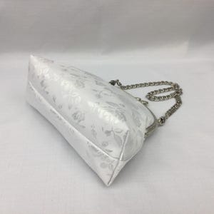 Puede incluir: Un bolso blanco con estampado floral y una correa de cadena plateada. El bolso tiene un cierre de broche.