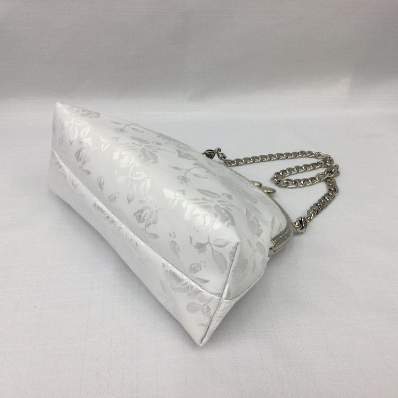 Puede incluir: Un bolso blanco con estampado floral y una correa de cadena plateada. El bolso tiene un cierre de broche.
