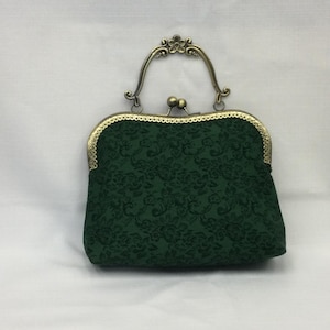 May include: A dark green, vintage-style handbag with a floral pattern. The bag features a decorative, ornate metal clasp and handle in a bronze finish. The bag's fabric has a subtle, embossed floral design.