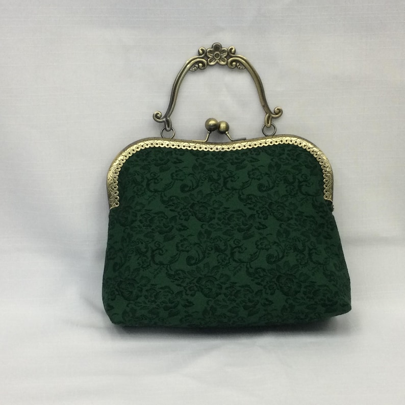 May include: A dark green, vintage-style handbag with a floral pattern. The bag features a decorative, ornate metal clasp and handle in a bronze finish. The bag's fabric has a subtle, embossed floral design.