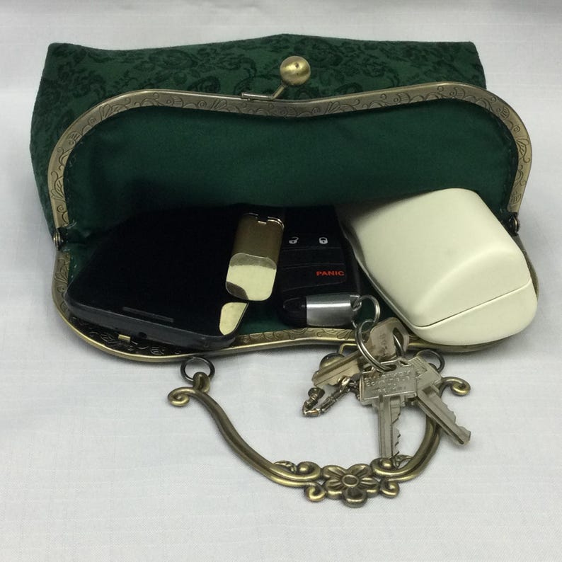 May include: Open green fabric clutch with a decorative brass frame and clasp. The interior reveals a black smartphone, car keys, a remote, and a gold-colored lighter. A decorative brass chain with keys hangs from the frame.