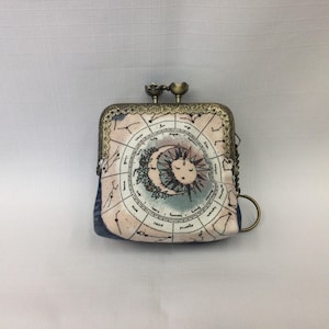 May include: A small, square coin purse with a metal clasp and a chain. The purse features a celestial design with a sun and moon in the center, surrounded by zodiac symbols. The color palette includes pink, blue, and cream.