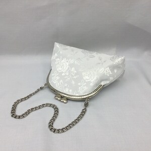 Puede incluir: Un bolso de sat&eacute;n blanco con un estampado floral y una correa de cadena plateada.