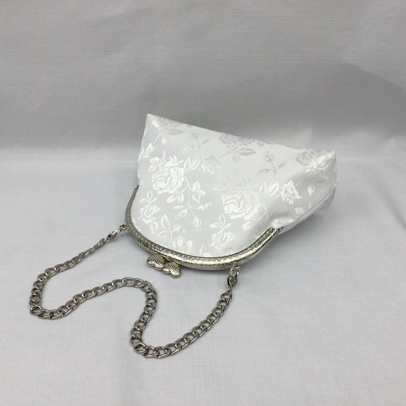 Puede incluir: Un bolso de sat&eacute;n blanco con un estampado floral y una correa de cadena plateada.