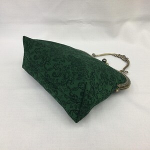 May include: A dark green clutch with a floral pattern. The bag has a metal clasp and handle with an antique brass finish. The fabric has a subtle, tone-on-tone design. The bag is rectangular in shape.
