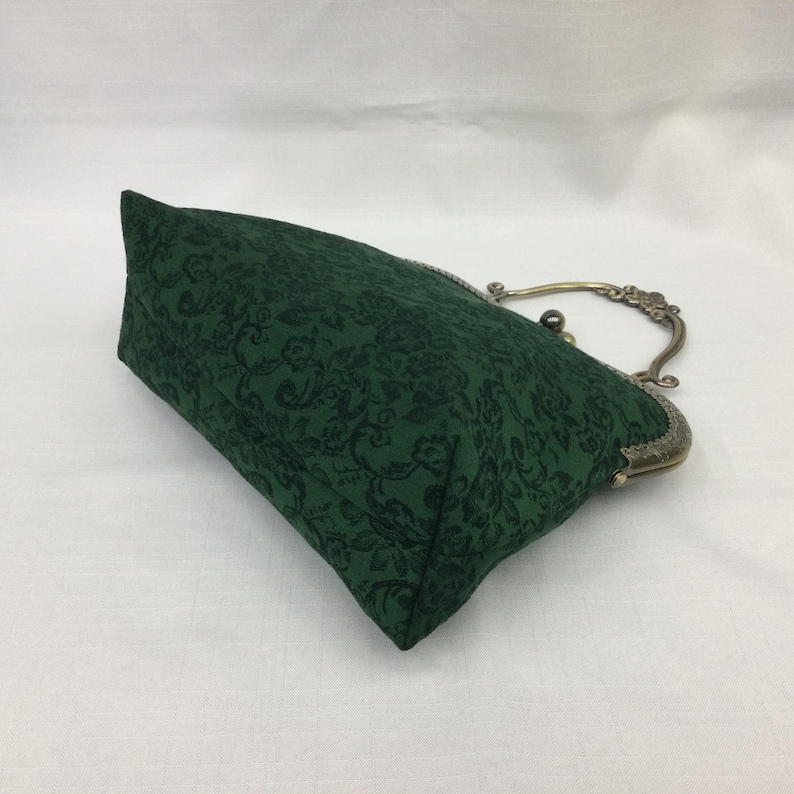 May include: A dark green clutch with a floral pattern. The bag has a metal clasp and handle with an antique brass finish. The fabric has a subtle, tone-on-tone design. The bag is rectangular in shape.