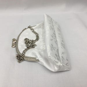 Puede incluir: Un bolso de mano de sat&eacute;n blanco con un estampado floral y una correa de cadena plateada. El bolso tiene un cierre de broche y un dise&ntilde;o en forma de coraz&oacute;n en la parte delantera.