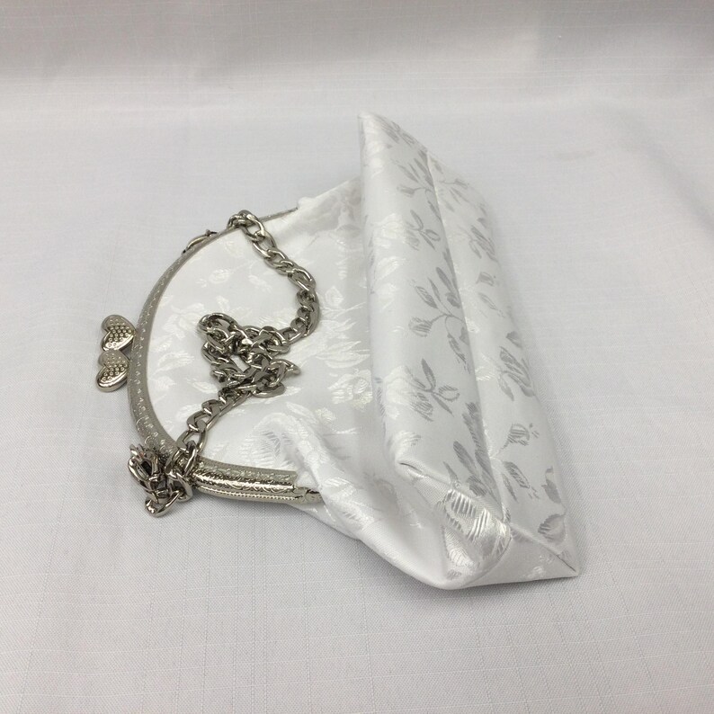 Puede incluir: Un bolso de mano de sat&eacute;n blanco con un estampado floral y una correa de cadena plateada. El bolso tiene un cierre de broche y un dise&ntilde;o en forma de coraz&oacute;n en la parte delantera.