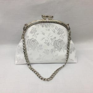 Puede incluir: Un bolso de mano blanco de sat&eacute;n con una correa de cadena plateada y un cierre en forma de coraz&oacute;n. El bolso tiene un patr&oacute;n floral.