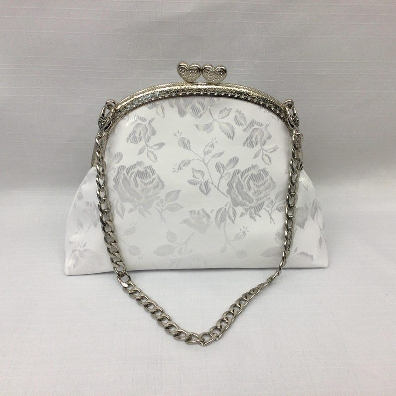 Puede incluir: Un bolso de mano blanco de sat&eacute;n con una correa de cadena plateada y un cierre en forma de coraz&oacute;n. El bolso tiene un patr&oacute;n floral.