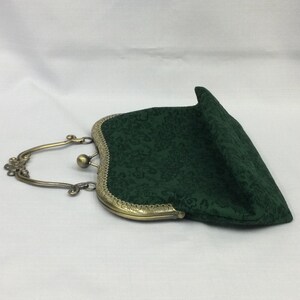 May include: A dark green fabric clutch with a floral pattern. The purse has a brass-colored metal frame and handle. The clasp is a small ball. The purse is open, revealing the interior.