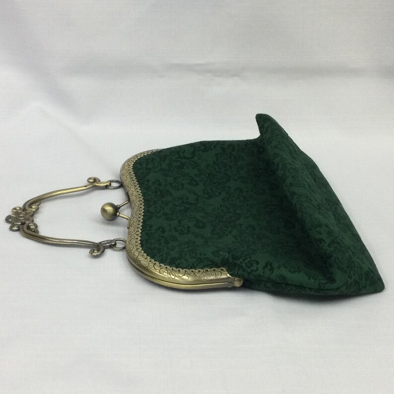 May include: A dark green fabric clutch with a floral pattern. The purse has a brass-colored metal frame and handle. The clasp is a small ball. The purse is open, revealing the interior.