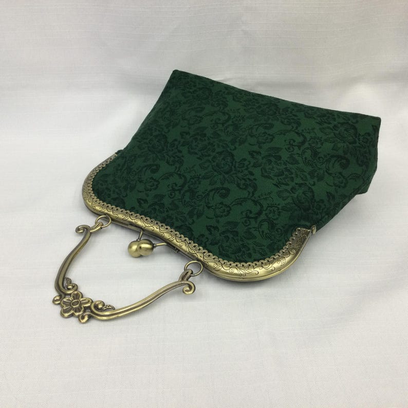May include: A dark green, floral-patterned clutch purse with a bronze-colored metal frame and handle. The handle features a decorative flower design. The purse is closed with a clasp.