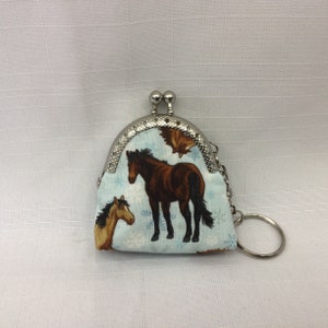 May include: A small coin purse with a silver clasp and a key ring. The purse is made of fabric with a light blue background and a pattern of brown horses and snowflakes. The clasp is silver.