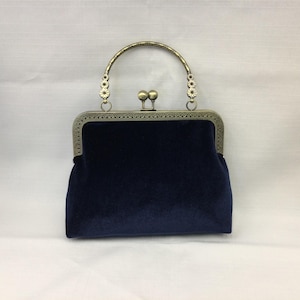 May include: A navy blue velvet handbag with a decorative metal frame and handle. The handle is arched and features small floral accents on each side. The clasp is a pair of small, round metal balls.