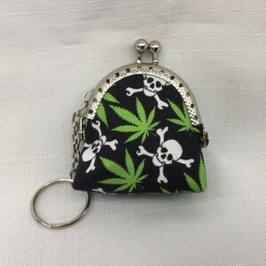 May include: A small black coin purse with a silver metal frame and a zipper closure. The purse is decorated with a green marijuana leaf and skull and crossbones pattern.