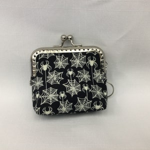 May include: A small black coin purse with a silver frame and a zip closure. The purse is decorated with a white spiderweb and spider pattern.