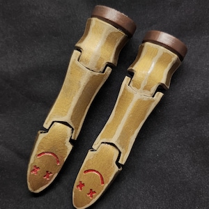 Può includere: Due estensioni per dita articolate in oro metallizzato con punte marroni. Ogni dito presenta una faccina sorridente rossa con occhi a forma di croce sulla punta. Le dita sono progettate per essere indossate e probabilmente fanno parte di un costume o cosplay.