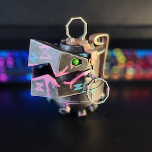 Könnte beinhalten: Eine 3D-gedruckte, schwarze, rosa und blaue Granate mit einem grünen Auge. Die Granate hat einen Metallring oben und einen Metallgriff an der Seite.