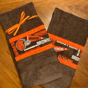 May include: Two brown kitchen towels with orange and black trim. The towels feature a Cleveland Browns logo and the words "Cleveland Browns" and "Dawg Pound".