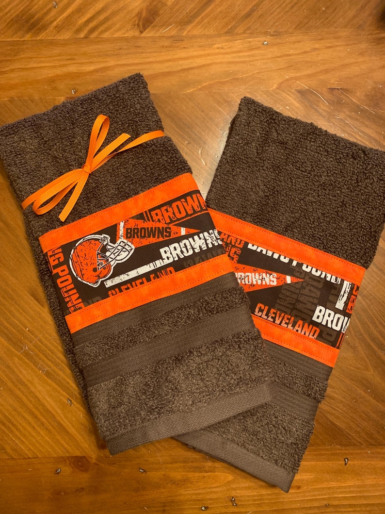 May include: Two brown kitchen towels with orange and black trim. The towels feature a Cleveland Browns logo and the words "Cleveland Browns" and "Dawg Pound".