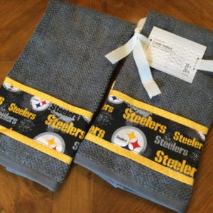 May include: Two gray kitchen towels with a yellow and black Steelers logo print. The towels have a white tag with the text "HAND TOWEL 16 x 26"