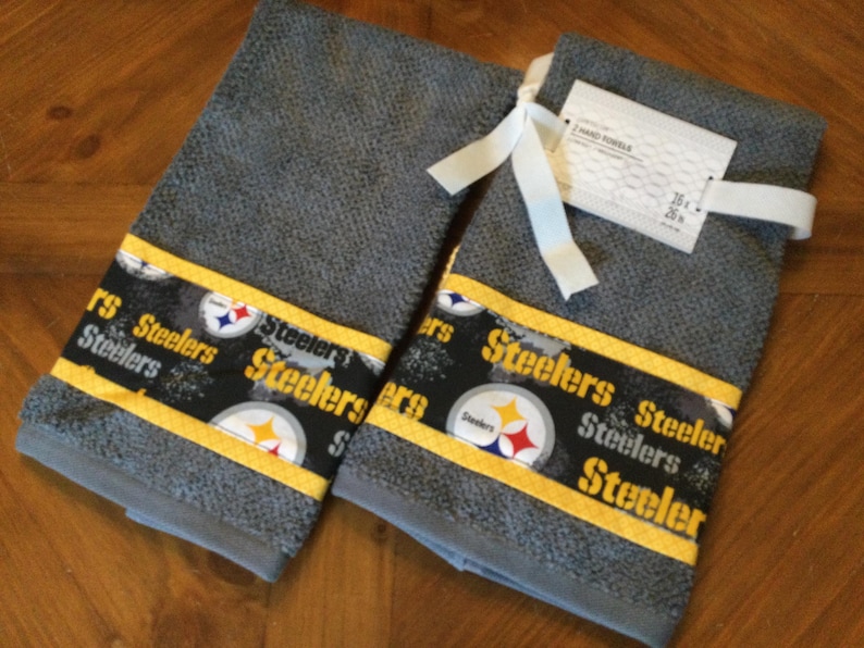May include: Two gray kitchen towels with a yellow and black Steelers logo print. The towels have a white tag with the text "HAND TOWEL 16 x 26"